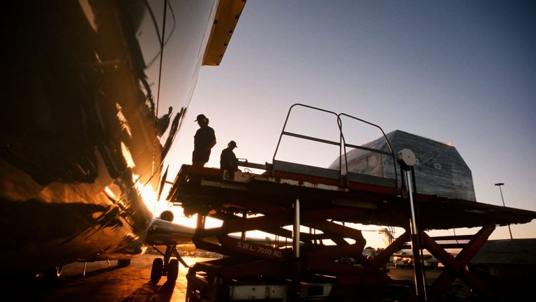 Workers are loading cargo freight onto an airplane using a platform lift during sunset.